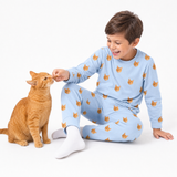 Child wearing a Sky Blue cat-print pajama set sitting on the floor and giving a treat to an orange cat.