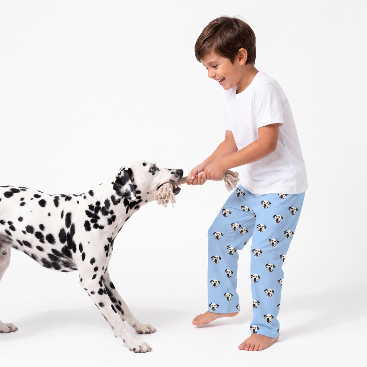 A child playing tug-of-war with a Dalmatian dog against a plain white studio background. The child is wearing a white t-shirt and light blue pajama pants printed with a repeating pattern of Dalmatian dog faces.