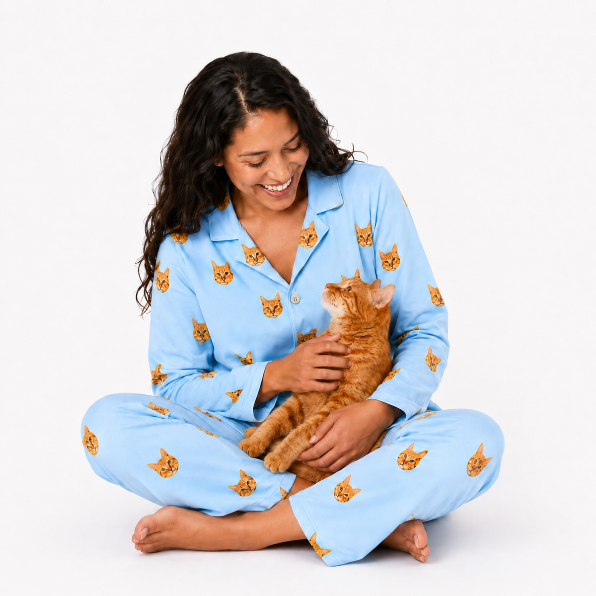 Woman wearing a Sky Blue cat-print pajama set sitting cross-legged and holding an orange cat.