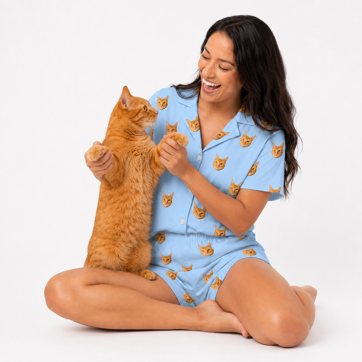 Woman in a sky blue pajama set with cat patterns sitting on the floor with an orange cat.