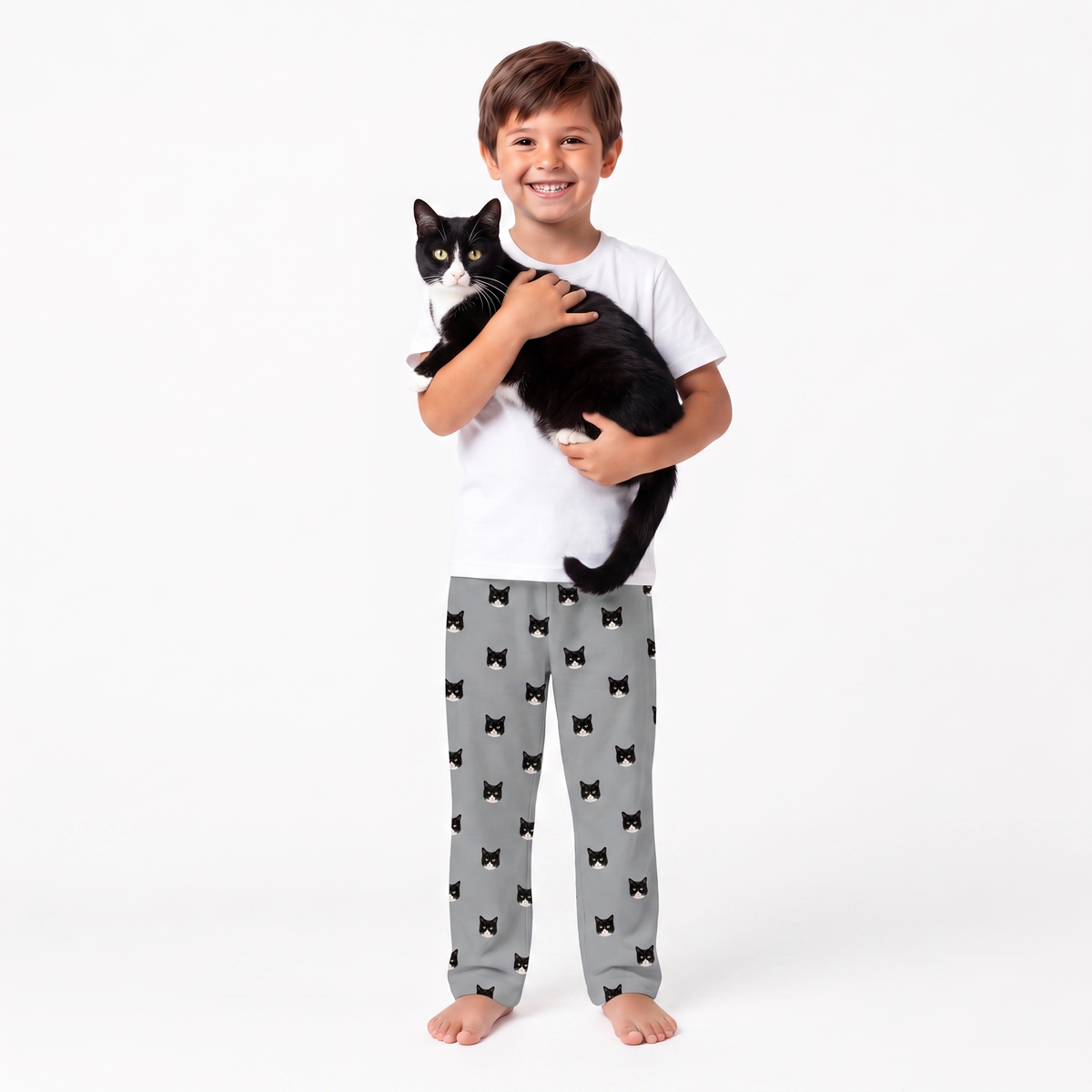 A young boy stands barefoot against a white studio background, holding a black cat. He is wearing a white t-shirt and slate gray pajama pants with a repeating black bow tie pattern.