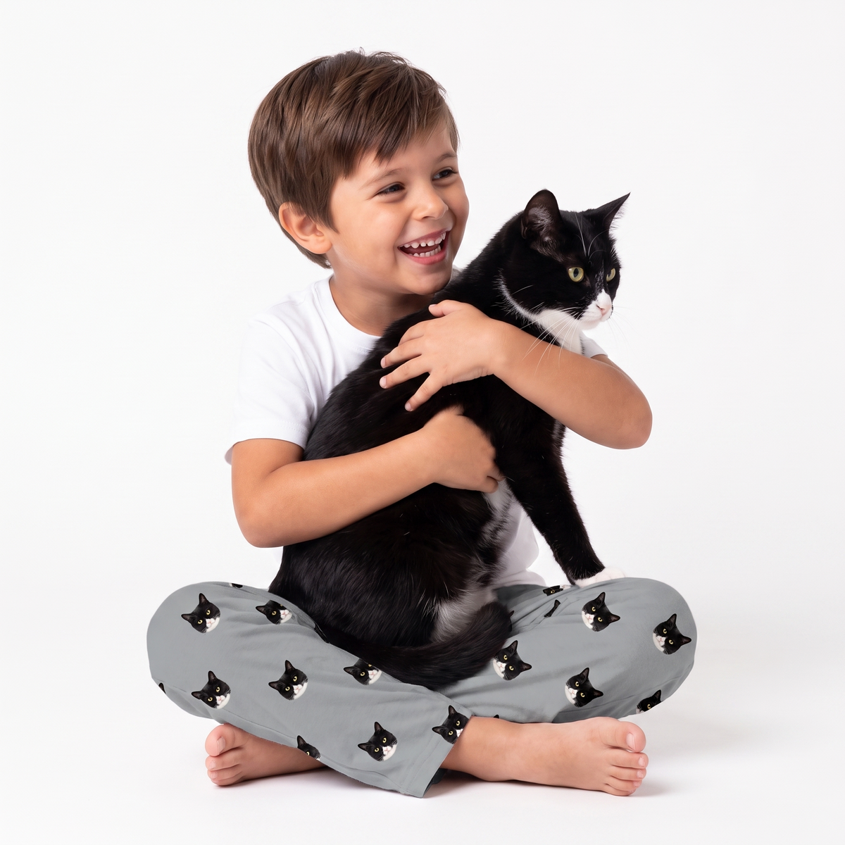 A young boy sitting cross-legged on a white studio background, smiling and hugging a black-and-white cat. He is wearing slate gray pajama pants covered in a repeating black-and-white cat face pattern.