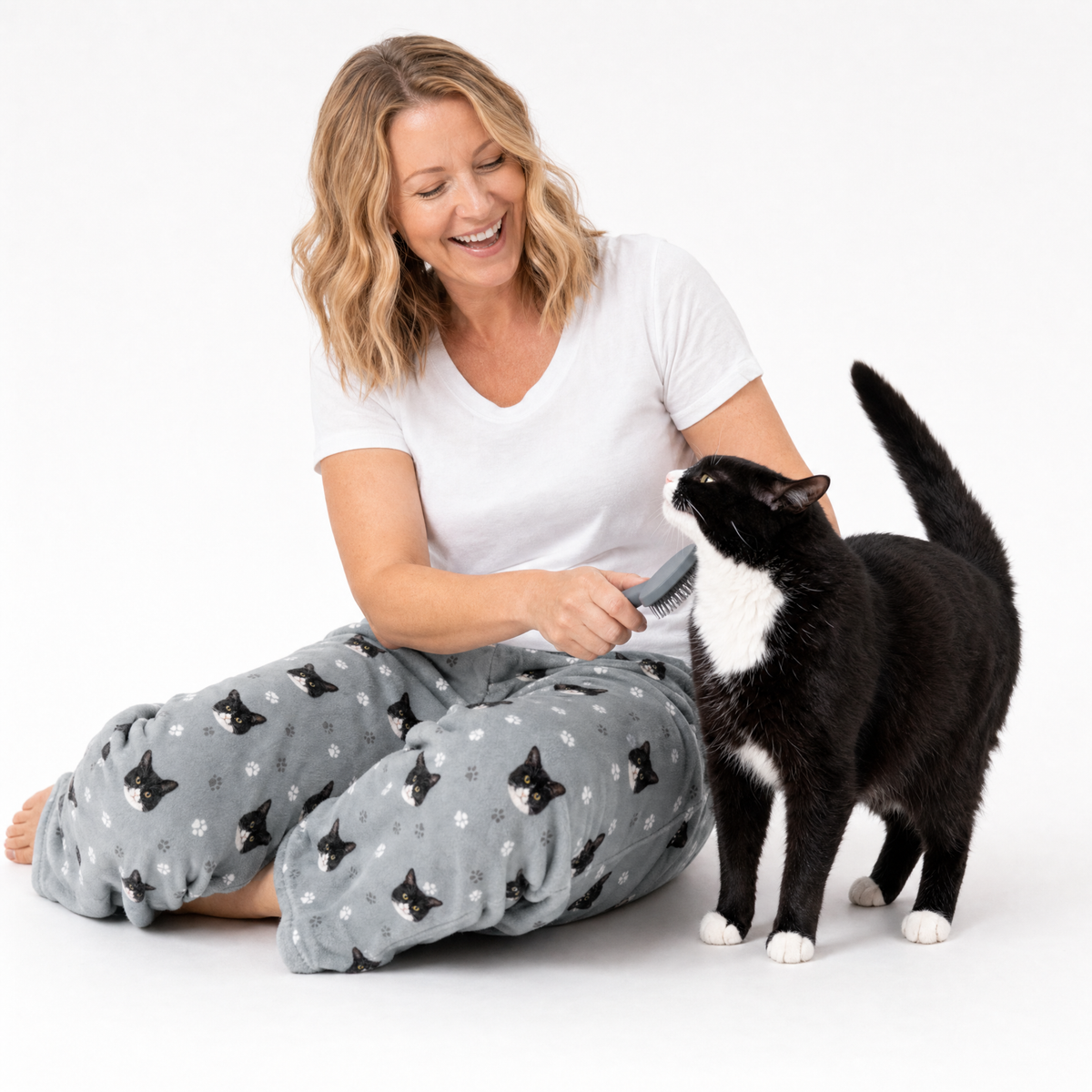 Woman wearing a white T-shirt and slate gray cat-print lounge pants while sitting on the floor and smiling as she brushes a black and white cat standing beside her.