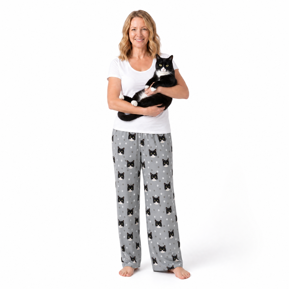Woman wearing a white top and gray cat patterned pants holding a black and white cat on a white background
