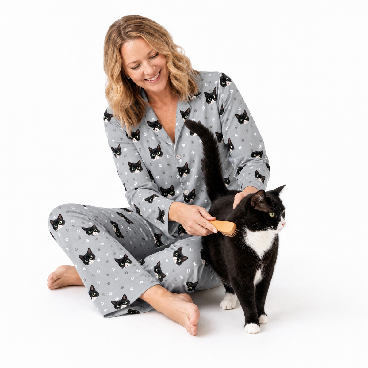 Woman in cat-themed pajamas brushing a black and white cat on a white background