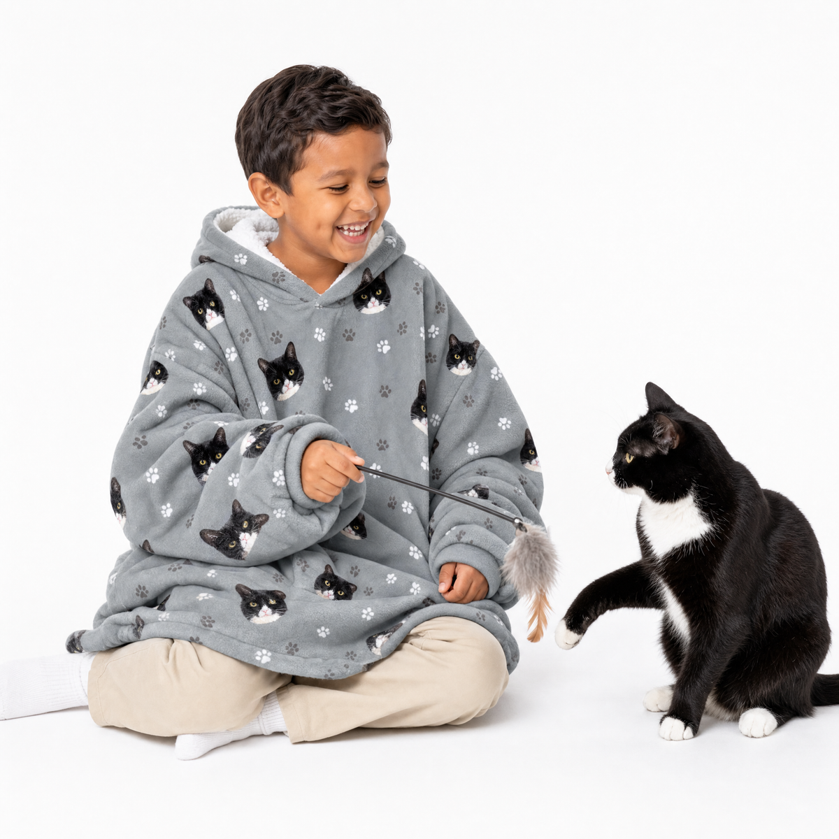 Child wearing a gray blanket hoodie with cat patterns, interacting with a black and white cat on a white background