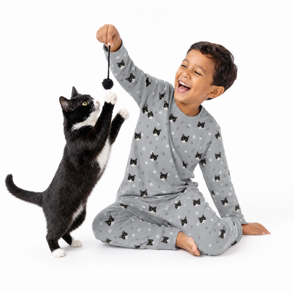 Child wearing a gray cat-print pajama set sitting on the floor while smiling and holding a dangling pom-pom toy as a black and white cat reaches up to play.