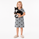 Young girl wearing a gray nightgown with black cat patterns, holding a black and white cat on a white background.