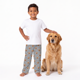A young boy and a golden retriever dog pose together on a white studio background. The boy wears a plain white t-shirt and slate gray pajama pants with a brown dog print, while the dog sits beside him.