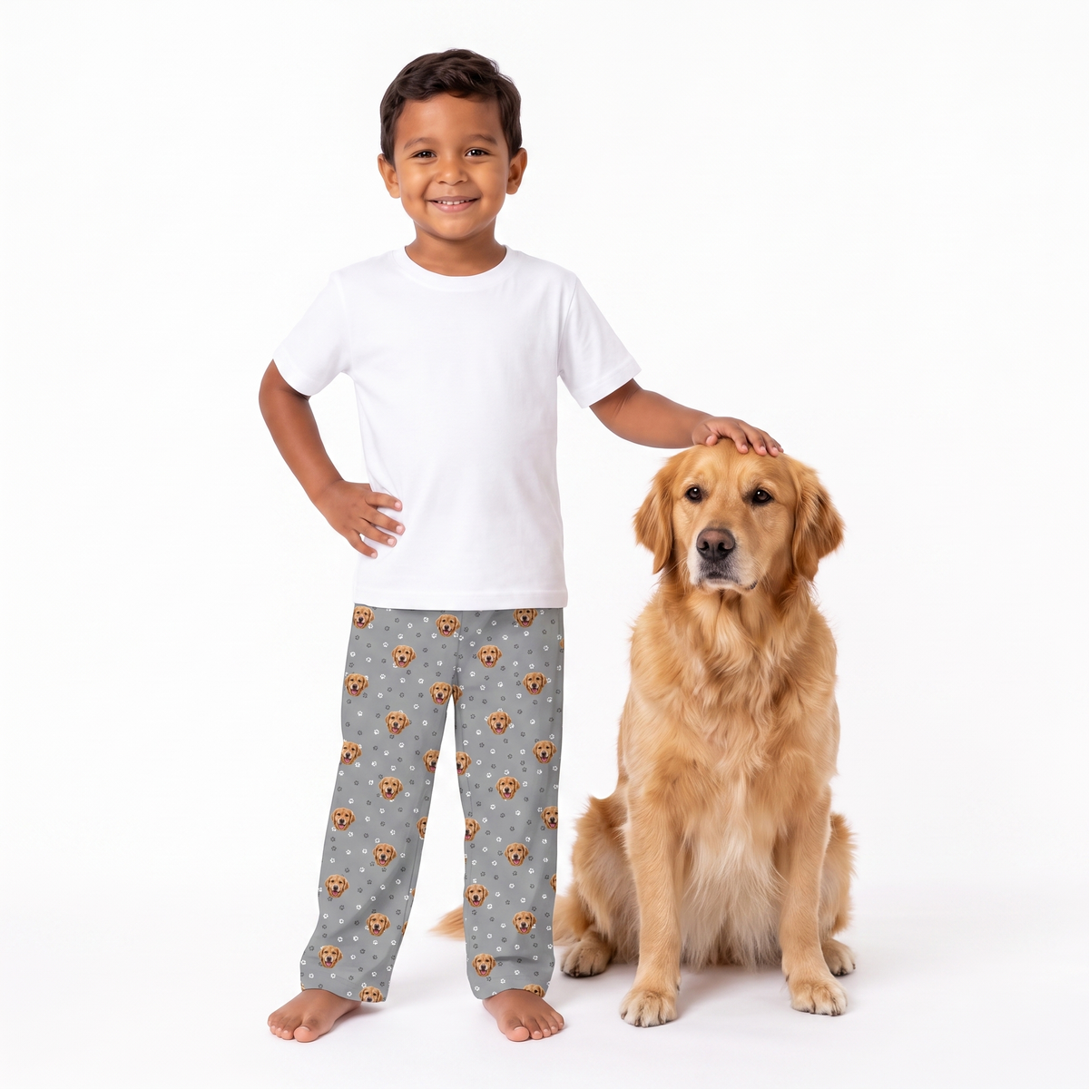 A young boy and a golden retriever dog pose together on a white studio background. The boy wears a plain white t-shirt and slate gray pajama pants with a brown dog print, while the dog sits beside him.