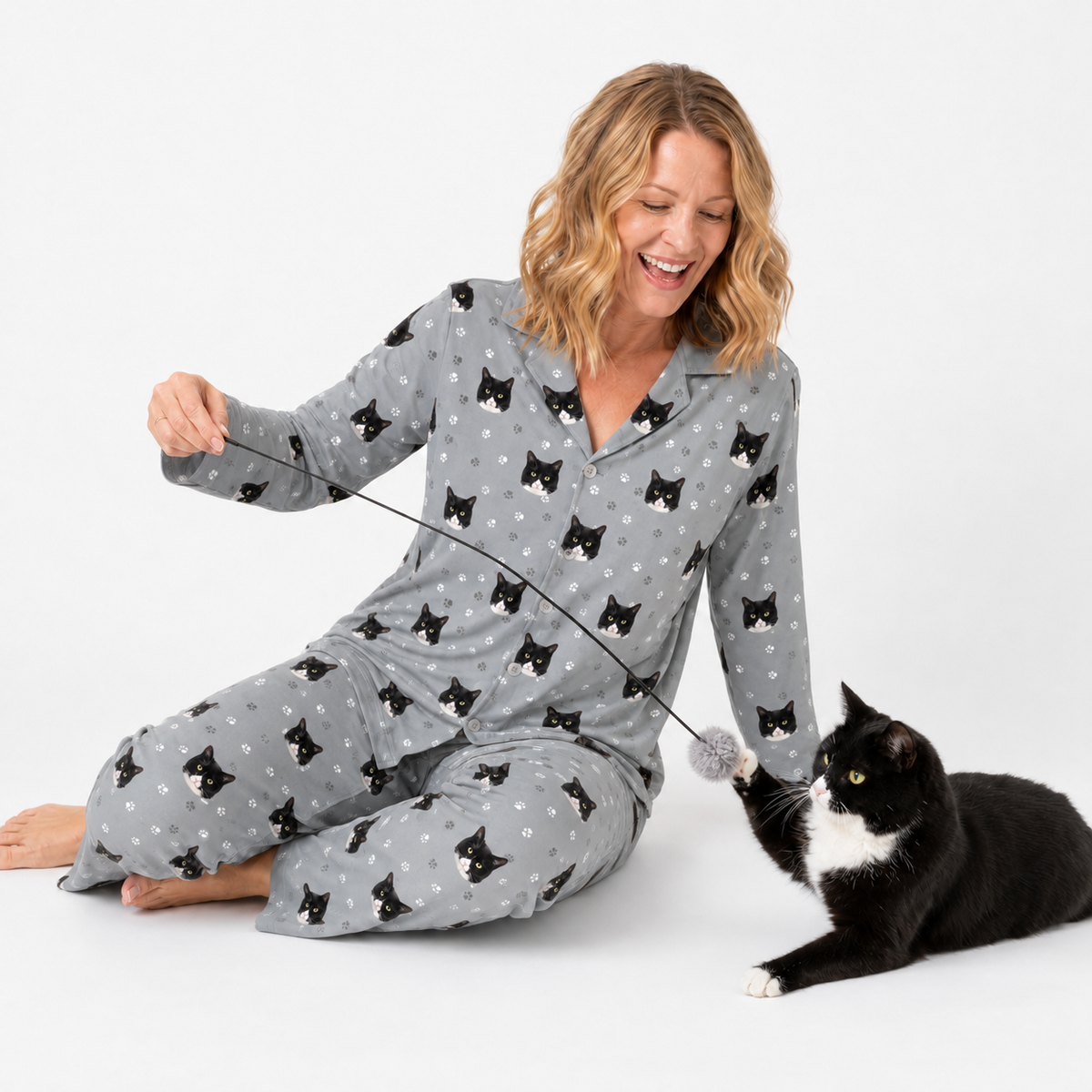 Woman wearing a Slate Gray Paws cat-print pajama set sitting on the floor and playing with a black and white cat.