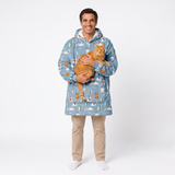 A man wearing a light blue hooded sweatshirt blanket with a winter forest print, holding an orange tabby cat, standing against a white studio background.