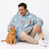 A man wearing a light blue hooded blanket sweater with a winter animal print sits on a white studio floor next to an orange tabby cat. The sweater features a pattern of polar bears, snow cats, pine trees, and snowflakes.
