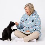 A woman sitting cross-legged on a white studio floor smiles while offering a treat to a black and white cat. She is wearing a light blue hooded blanket pajama cover with a winter animal print, including husky dogs, snowmen, and pine trees.
