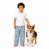 A young boy wearing a white t-shirt and light blue pajama pants with a dog print stands next to a corgi on a solid white background.