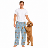 A man wearing light blue pajama pants with a woodland holiday print stands next to a small brown dog on a white studio background. The pants feature a pattern of pine trees, snow, pine cones, and brown pine trees.