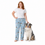 A woman stands barefoot next to a dog wearing light blue pajama pants with a Christmas-themed print of Santa Claus, snowman, pine trees, and snowflakes. The woman is wearing a plain white t-shirt, and the dog is wearing a fluffy white, gray, and brown winter coat.