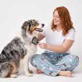 A woman with red hair sits on the floor brushing a long-haired dog with a black grooming brush. She is wearing a white t-shirt and light blue pajama pants with a dog print, against a plain white studio background.