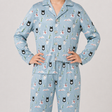 A person wearing a light blue long-sleeve pajama set with a festive Christmas print featuring black cats, snowmen, pine trees, and snowflakes, posed against a neutral light gray background.