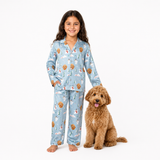A young girl stands barefoot next to a brown poodle dog wearing a light blue pajama set with a Christmas dog print, including Santa Claus, snowman, and golden retriever motifs, against a plain white background.