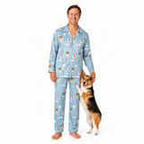A man wearing light blue pajamas with a dog print stands barefoot against a white studio background, holding a corgi dog that is standing on its hind legs to pet him.