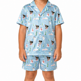 A child wearing a light blue short-sleeve pajama set with a repeating print of dogs, snowmen, Christmas trees, and mountains, shown against a plain white background.