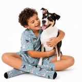 A smiling child wearing light blue pajamas with a dog print sits cross-legged on a white studio background, hugging a black, white, and tan dog. The pajamas and matching socks feature a repeating dog-and-tree pattern.