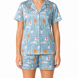 A woman modeling a light blue short-sleeve pajama set with a repeating print of golden retriever dogs, snowmen, snowy mountains, and evergreen trees on a white background.