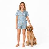 A woman wearing a light blue pajama set with a whimsical cartoon print stands barefoot next to a golden retriever dog on a solid white background.