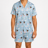 A man wearing a light blue short-sleeve pajama shirt and matching shorts with a repeating Christmas-themed print of dogs, snowmen, and trees, photographed against a plain light gray studio background.