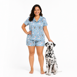 A woman wearing a light blue pajama set with a dog-themed print stands barefoot next to a black-and-white Dalmatian on a plain white studio background.