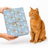 A ginger orange cat sits beside a wrapped gift box held by two hands against a clean white background. The gift is wrapped in light blue paper with a repeating pattern of white cats, orange fish, and small holiday motifs, tied with a brown string.