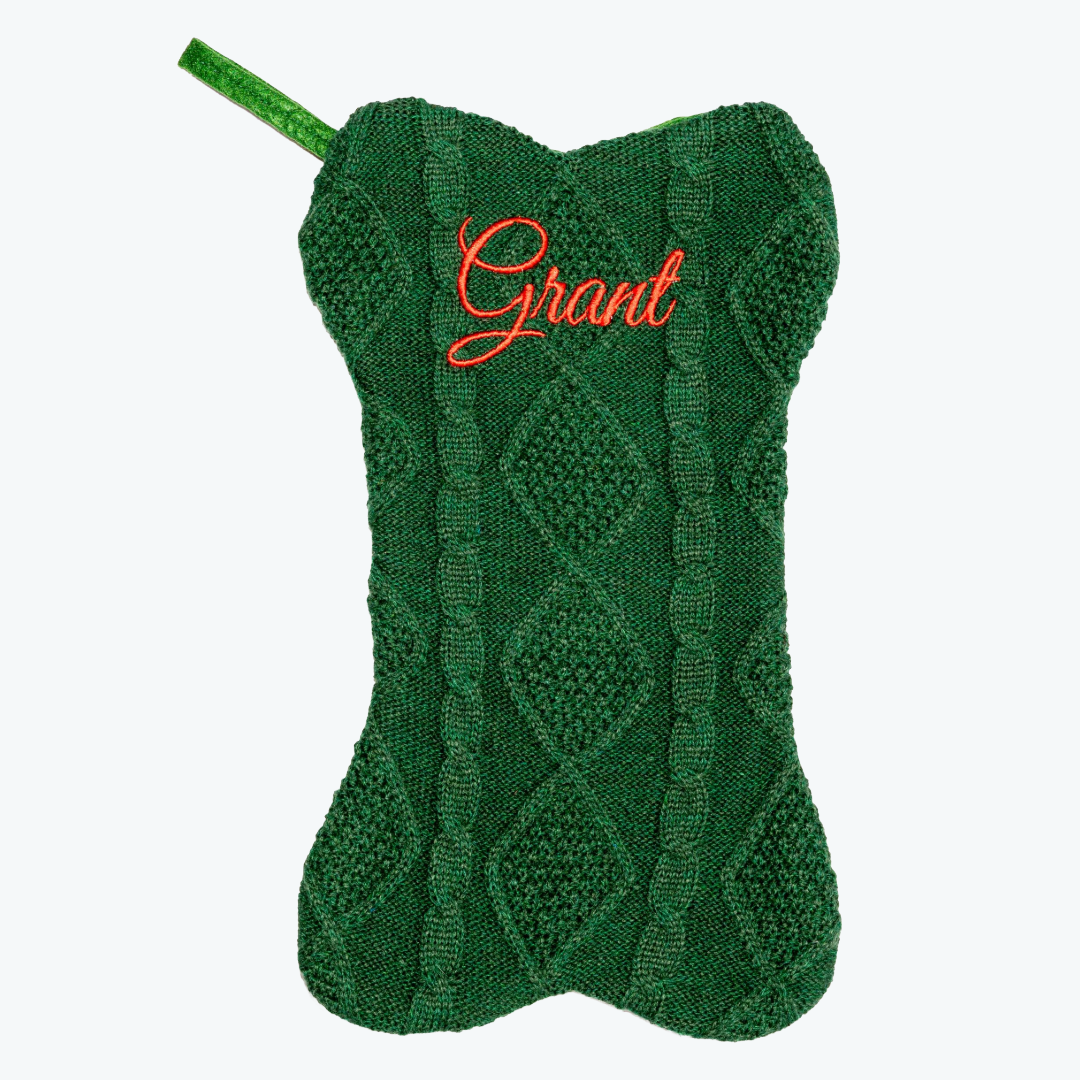 Green knitted dog-bone–shaped Christmas stocking with a cable knit pattern and the name ‘Grant’ embroidered in red script at the top, featuring a matching green hanging loop.