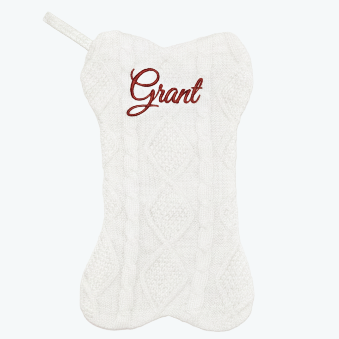 White knitted dog-bone–shaped Christmas stocking with a cable knit pattern and the name ‘Grant’ embroidered in red script at the top, with a matching hanging loop on the side.