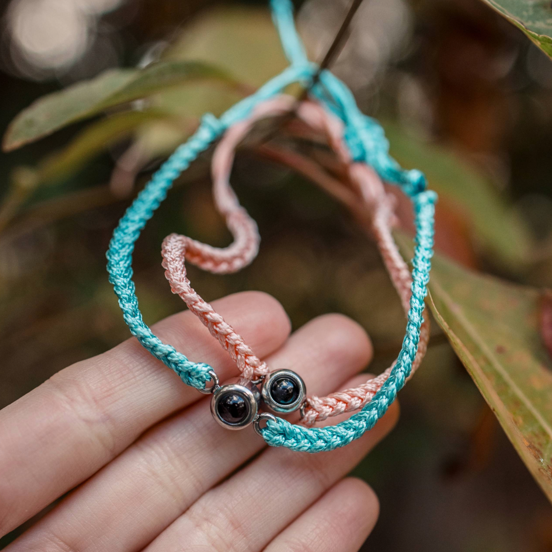 Pink and blue braided bracelets with a black bead charm held between fingers.
