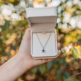 Hand holding open jewelry box with silver necklace outdoors, sunlit bokeh background.