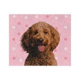 A blanket with a pink background and paw prints, featuring a centered, smiling portrait of a dog.