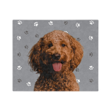 A blanket with a gray background and paw prints, featuring a centered, smiling portrait of a dog.