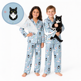 Two children wearing matching light blue pajamas with a black-and-white cat print, standing barefoot on a white studio background. One child holds a black-and-white cat, and a circular inset at the upper left shows a close-up of the cat motif printed on the pajamas.
