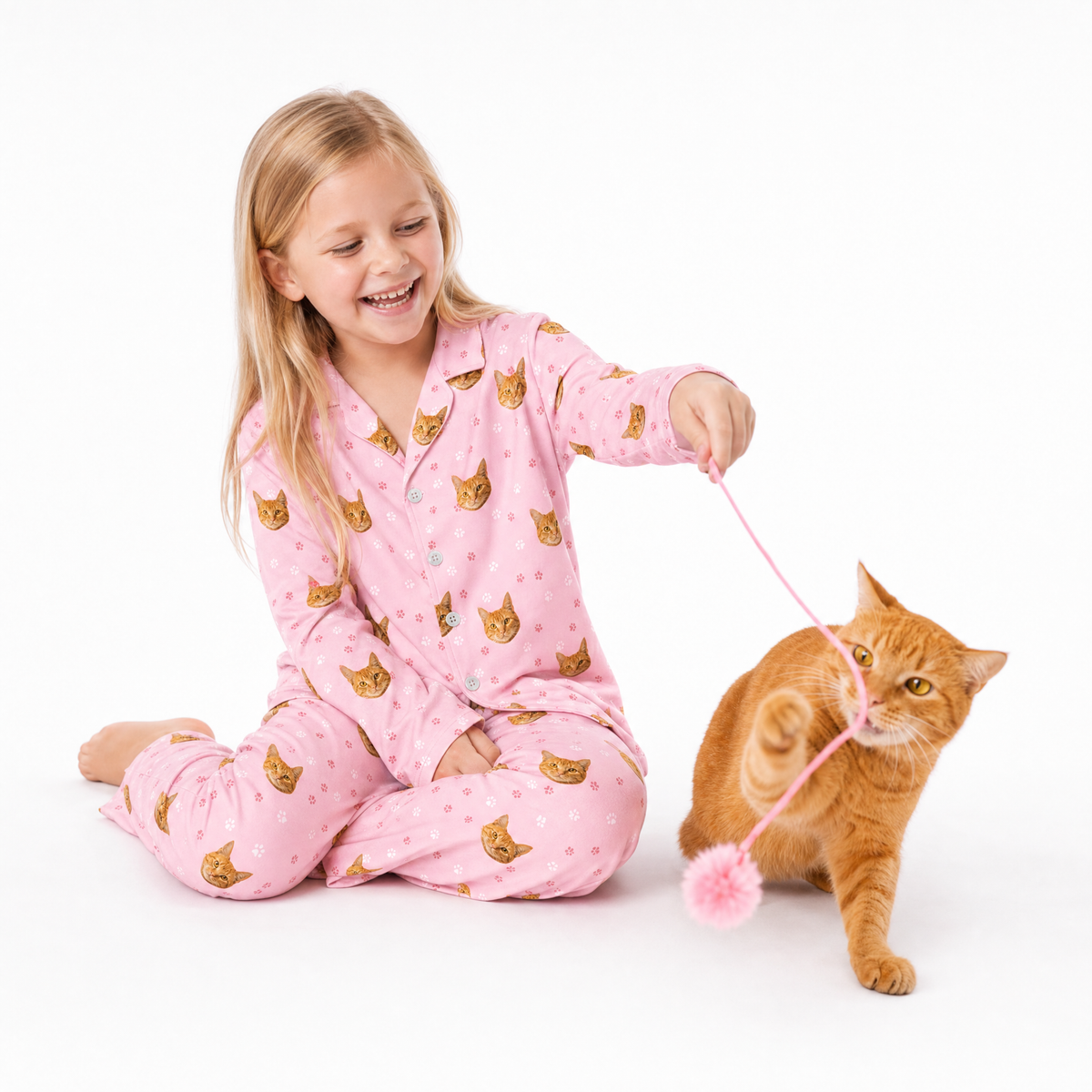 Child wearing a Light Pink Paws cat-print pajama set sitting on the floor and playing with an orange cat using a pink toy.