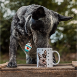 A black and gray speckled dog leans down to sniff a white ceramic mug of itself.