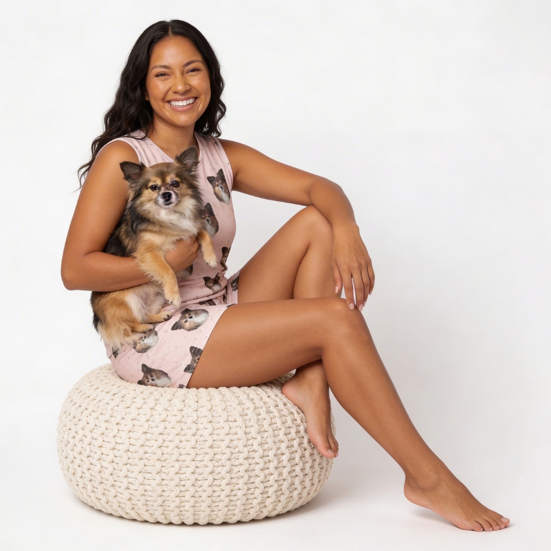 Woman wearing tank top pajamas set and sitting on a woven ottoman holding a small dog, wearing a pink dress with cat print.